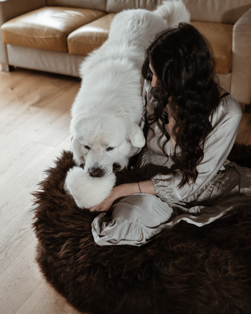 Woman sitting on a dark sheepskin dog bed, gently holding a white heart-shaped sheepskin toy as a large white dog engages with it—natural, non-toxic play from Mellow Concept Store.
