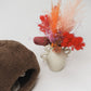 Small vase with colorful dried flowers next to a brown sheepskin pet bed with cat hiding inside, a white background | Natural, non‑toxic pet nest for cats and small dogs | Cozy, handmade pet furniture, natural wool and sheepskin

