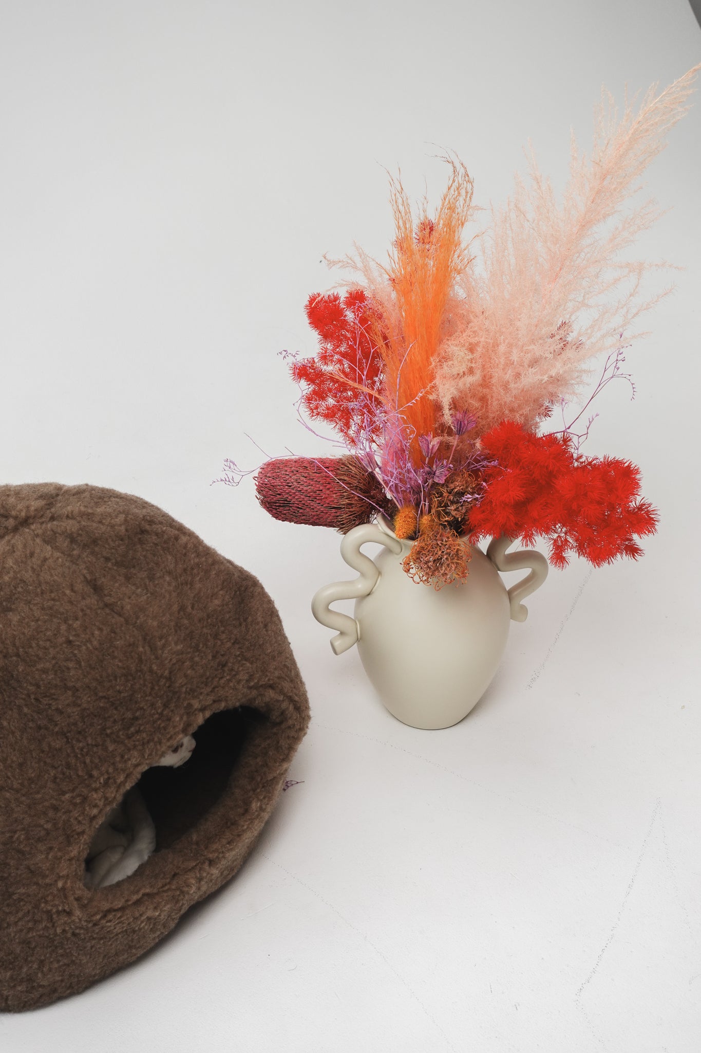 Small vase with colorful dried flowers next to a brown sheepskin pet bed with cat hiding inside, a white background | Natural, non‑toxic pet nest for cats and small dogs | Cozy, handmade pet furniture, natural wool and sheepskin

