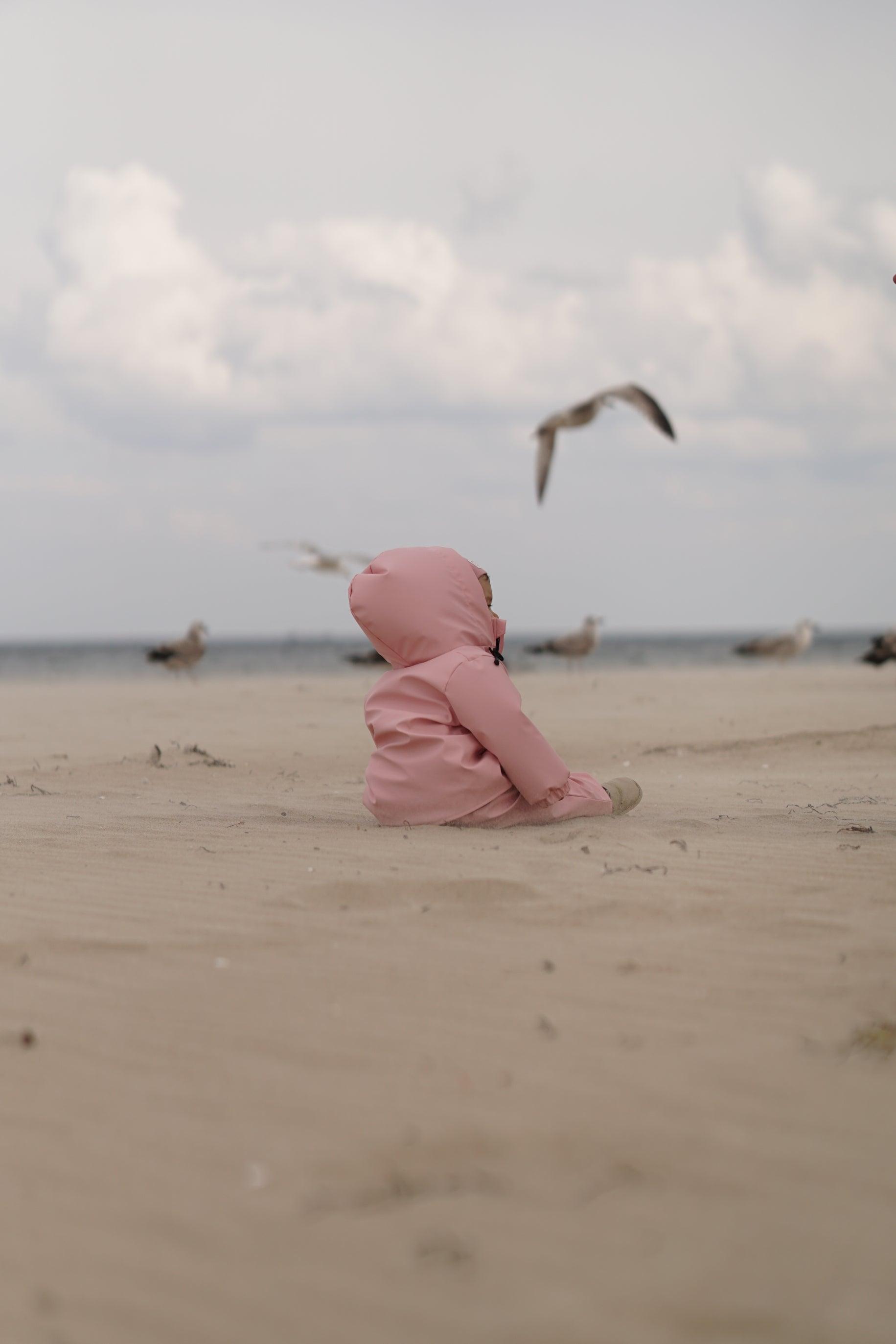 A child in the MellowConceptStore Waterproof Baby/Kid Clothing Set - Rosa sits alone on a sandy beach, gazing at a seagull flying by, with more seagulls and a cloudy sky in the background.