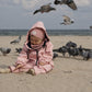 A child in a pink jacket with adjustable suspenders sits on the sand surrounded by pigeons, with some birds in flight near them wearing the Waterproof Baby/Kid Clothing Set - Rosa from MellowConceptStore.