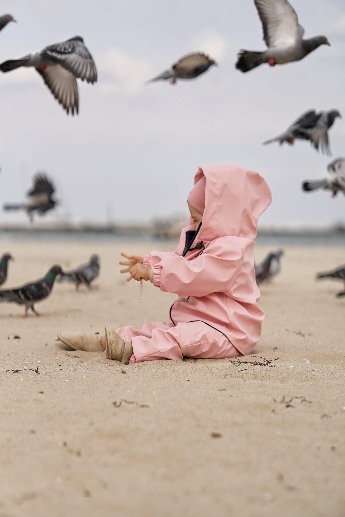 A child in a Waterproof Baby/Kid Clothing Set - Rosa from MellowConceptStore is sitting on a sandy beach surrounded by pigeons in flight and on the ground.
